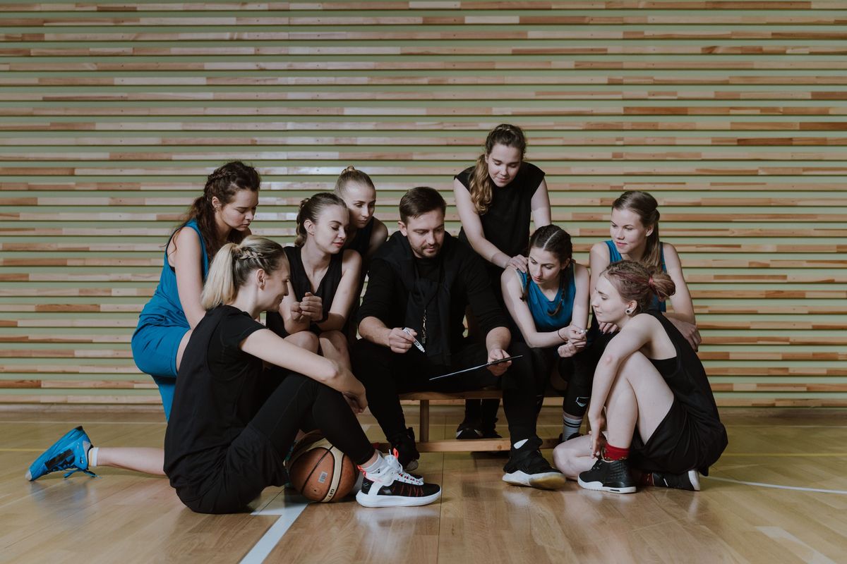 A girls basketball team huddled around their coach as he explains plays on a clipboard in a gym