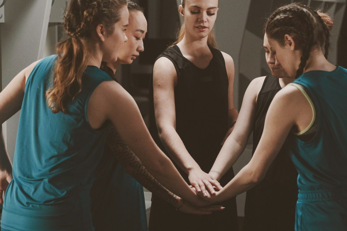Five women basketball players in a locker room huddle, hands stacked together