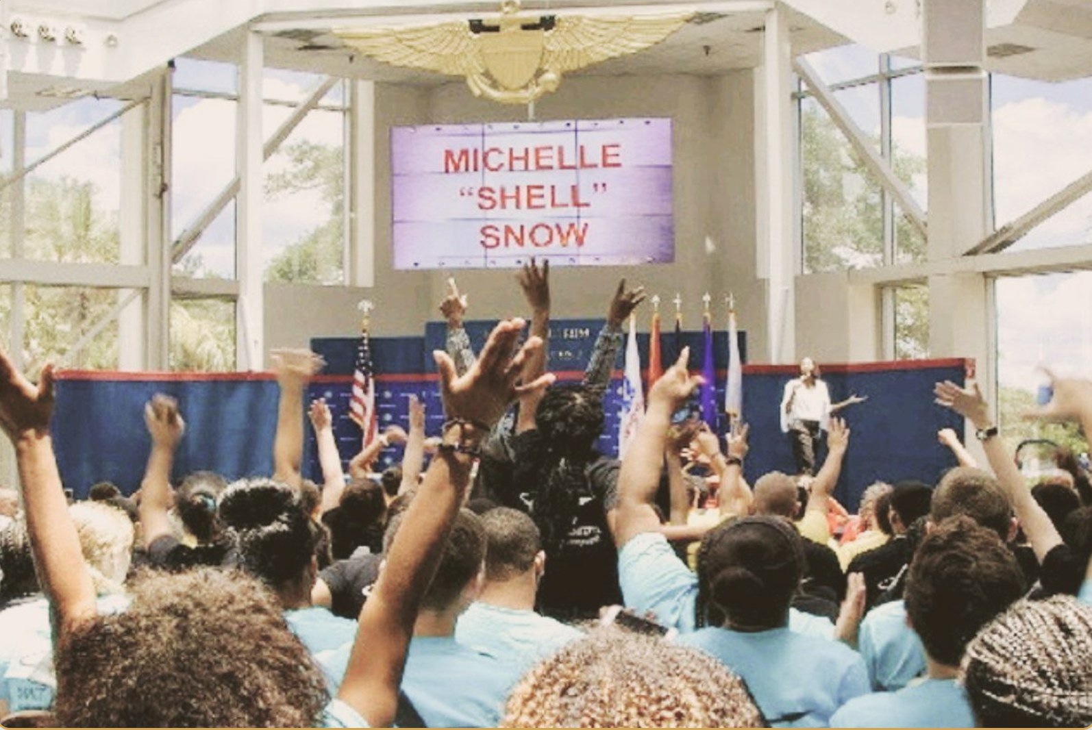 Michelle Snow speaking on stage to a large youth audience with hands raised at the National Naval Aviation Museum
