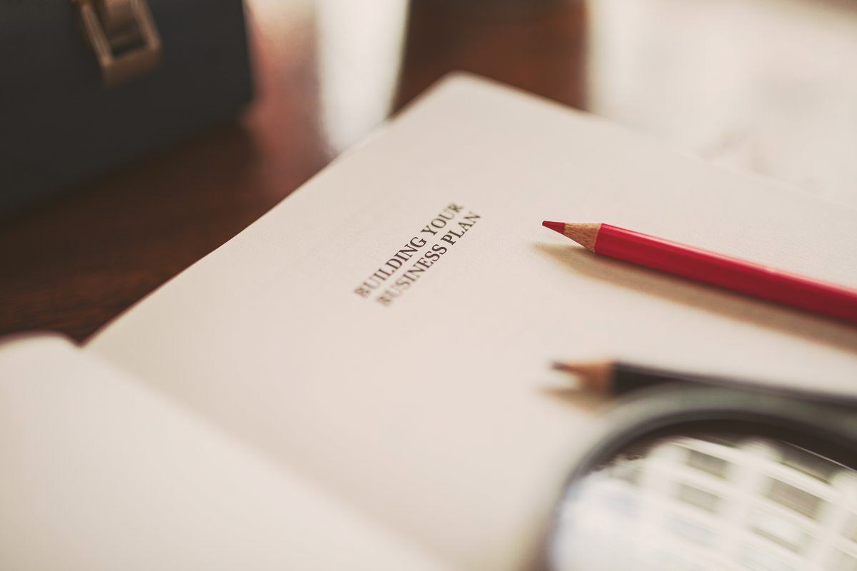 An open business plan notebook on a desk with pencils and a magnifying glass